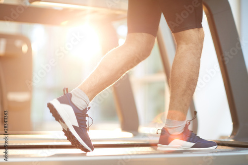 Sports background of muscular male legs running on treadmill in gym lit by sunlight, copy space