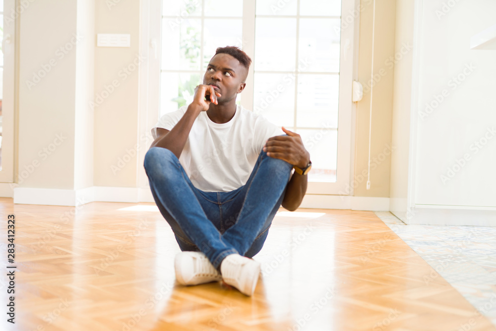 Handsome african american man sitting on the floor at home with hand on chin thinking about question, pensive expression. Smiling with thoughtful face. Doubt concept. Handsome african american man sitting on the floor at home with hand on chin thinking about question, pensive expression. Smiling with thoughtful face. Doubt concept.