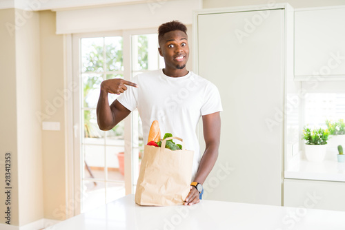 African american man holding paper bag full of fresh groceries with surprise face pointing finger to himself