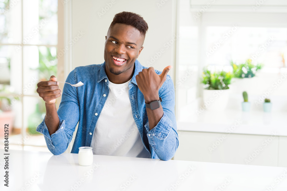 African american man eating healthy natural yogurt with a spoon pointing and showing with thumb up to the side with happy face smiling African american man eating healthy natural yogurt with a spoon pointing and showing with thumb up to the side with happy face smiling