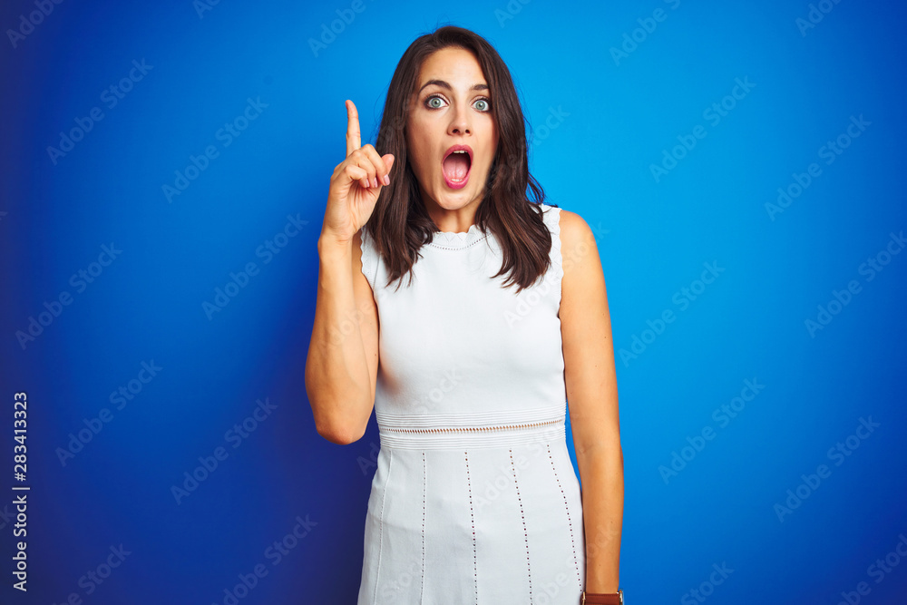 Young business woman wearing white elegant dress standing over blue isolated background pointing finger up with successful idea. Exited and happy. Number one. Young business woman wearing white elegant dress standing over blue isolated background pointing finger up with successful idea. Exited and happy. Number one.