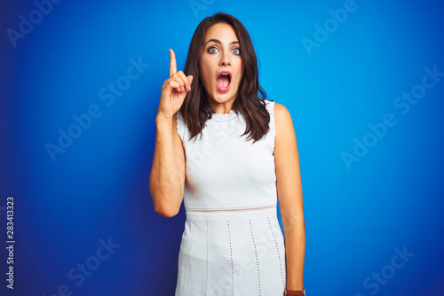 Young business woman wearing white elegant dress standing over blue isolated background pointing finger up with successful idea. Exited and happy. Number one.