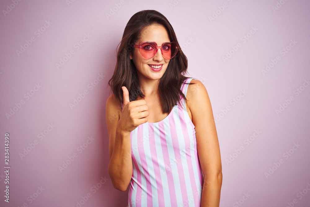 Beautiful woman wearing striped pink swimsuit and heart shaped sunglasses over pink background doing happy thumbs up gesture with hand. Approving expression looking at the camera with showing success.