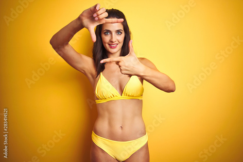 Beautiful woman wearing yellow bikini on summer over isolated yellow background smiling making frame with hands and fingers with happy face. Creativity and photography concept. © Krakenimages.com