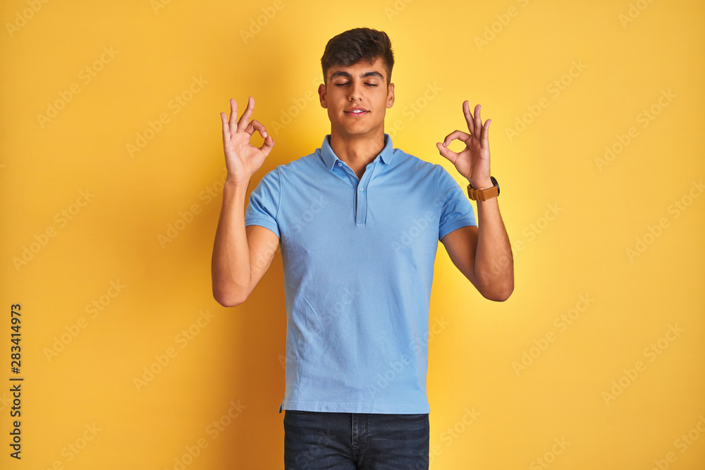 Young indian man wearing blue polo standing over isolated yellow background relax and smiling with eyes closed doing meditation gesture with fingers. Yoga concept. Young indian man wearing blue polo standing over isolated yellow background relax and smiling with eyes closed doing meditation gesture with fingers. Yoga concept.