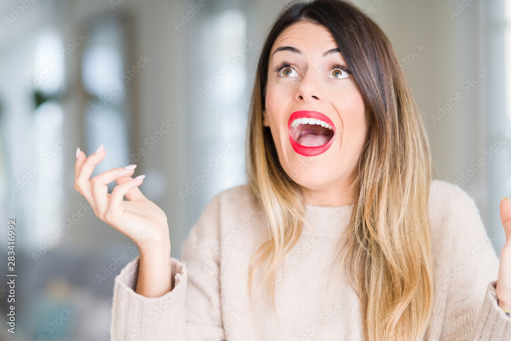 Young beautiful woman wearing winter sweater at home crazy and mad shouting and yelling with aggressive expression and arms raised. Frustration concept. Young beautiful woman wearing winter sweater at home crazy and mad shouting and yelling with aggressive expression and arms raised. Frustration concept.