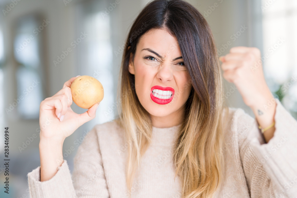Young beautiful woman holding fresh potato at home annoyed and frustrated shouting with anger, crazy and yelling with raised hand, anger concept Young beautiful woman holding fresh potato at home annoyed and frustrated shouting with anger, crazy and yelling with raised hand, anger concept