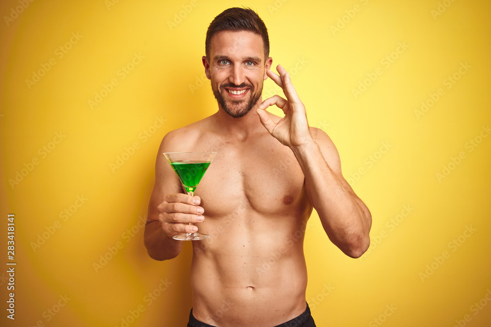 Young handsome shirtless man drinking a summer cocktail over isolated yellow background doing ok sign with fingers, excellent symbol