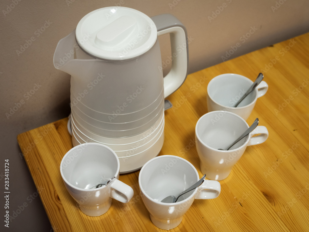 White electric kettle and four white ceramic mugs with teaspoons on wooden table White electric kettle and four white ceramic mugs with teaspoons on wooden table