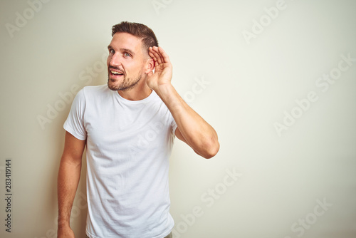 Young handsome man wearing casual white t-shirt over isolated background smiling with hand over ear listening an hearing to rumor or gossip. Deafness concept.