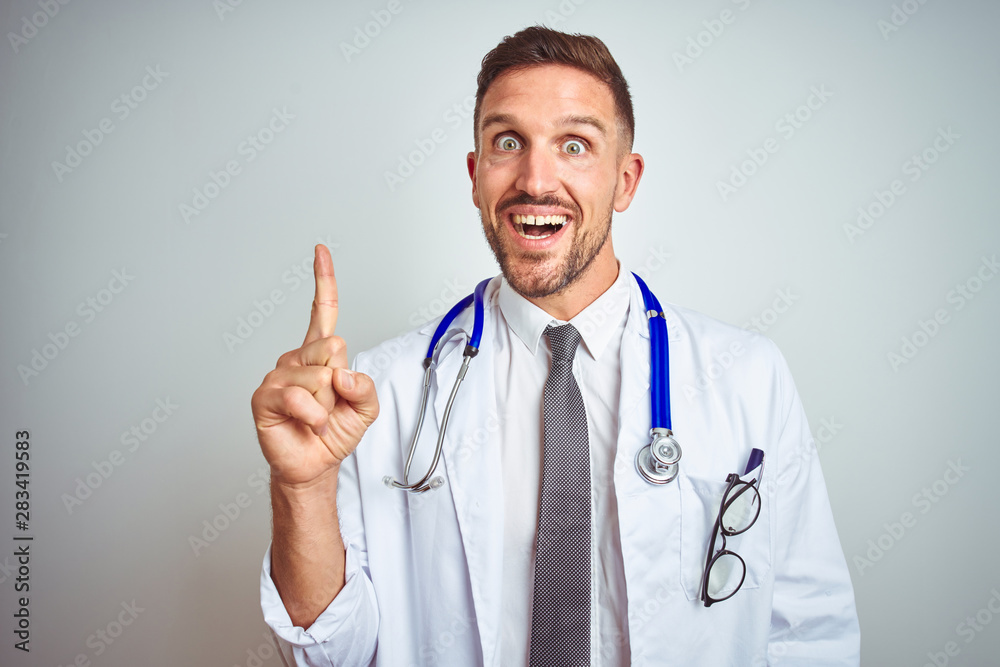 Young handsome doctor man wearing white professional coat over isolated background pointing finger up with successful idea. Exited and happy. Number one. Young handsome doctor man wearing white professional coat over isolated background pointing finger up with successful idea. Exited and happy. Number one.