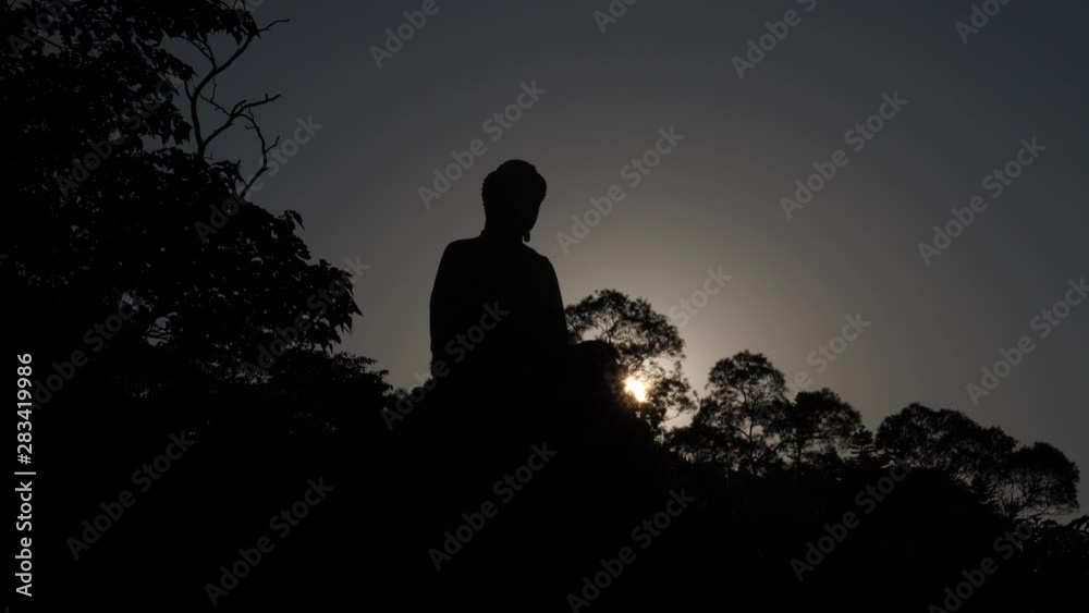 Silhouetted low angle shot of Tian Tan Buddha at Lantau Island, Hong Kong. Large bronze statue shadow against dark sky at evening time. Black foliage of trees surround famous religious site Silhouetted low angle shot of Tian Tan Buddha at Lantau Island, Hong Kong. Large bronze statue shadow against dark sky at evening time. Black foliage of trees surround famous religious site