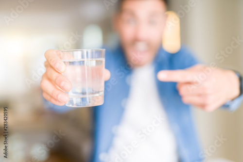 Young handsome man drinking a glass of water at home very happy pointing with hand and finger