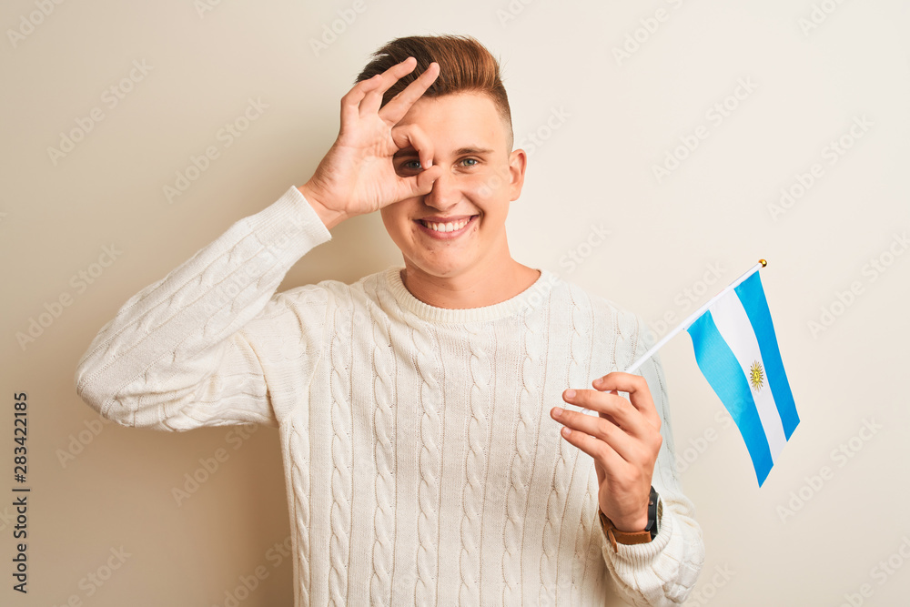 Young handsome man holding Argentinian Argentina flag over isolated white background with happy face smiling doing ok sign with hand on eye looking through fingers Young handsome man holding Argentinian Argentina flag over isolated white background with happy face smiling doing ok sign with hand on eye looking through fingers