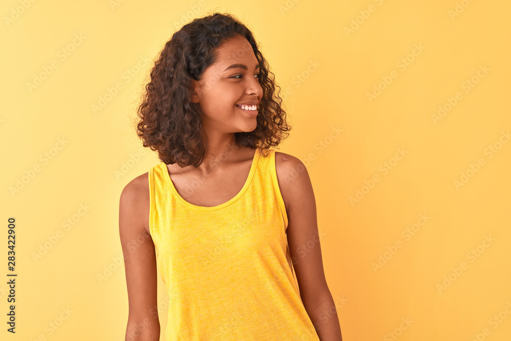Young brazilian woman wearing t-shirt standing over isolated yellow background looking away to side with smile on face, natural expression. Laughing confident. Young brazilian woman wearing t-shirt standing over isolated yellow background looking away to side with smile on face, natural expression. Laughing confident.