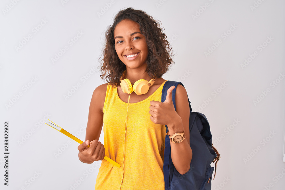 Brazilian student woman wearing backpack holding notebook over isolated white background happy with big smile doing ok sign, thumb up with fingers, excellent sign - obrazy, fototapety, plakaty 