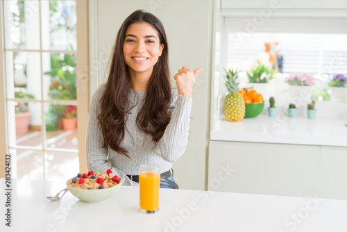 Young woman eating healthy breakfast in the morning smiling with happy face looking and pointing to the side with thumb up.