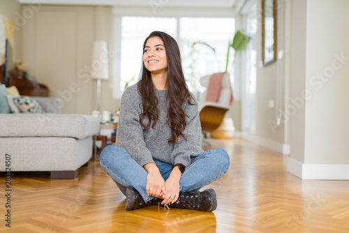 Young beautiful woman sitting on the floor at home looking away to side with smile on face, natural expression. Laughing confident. © Krakenimages.com