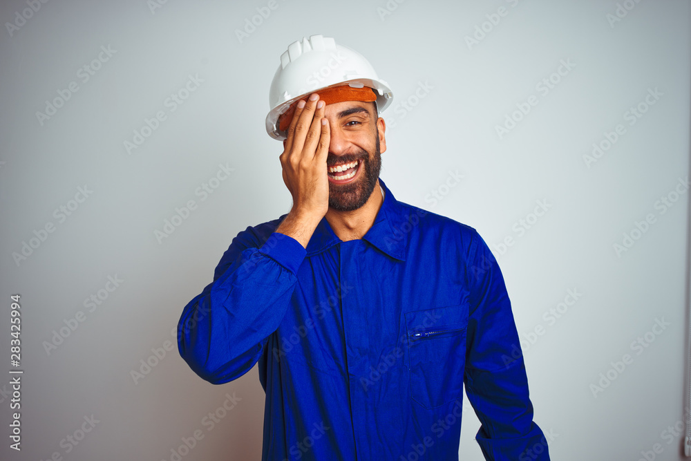 Handsome indian worker man wearing uniform and helmet over isolated white background covering one eye with hand, confident smile on face and surprise emotion.