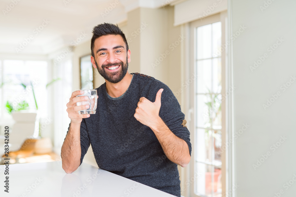 Handsome hispanic man drinking a fresh glass of water happy with big smile doing ok sign, thumb up with fingers, excellent sign Handsome hispanic man drinking a fresh glass of water happy with big smile doing ok sign, thumb up with fingers, excellent sign