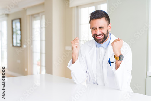 Handsome hispanic doctor or therapist man wearing medical coat at the clinic excited for success with arms raised celebrating victory smiling. Winner concept.