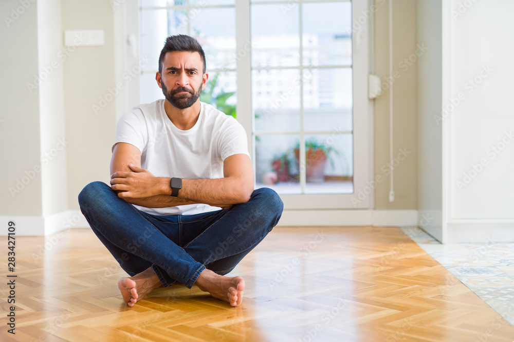 Handsome hispanic man wearing casual t-shirt sitting on the floor at home skeptic and nervous, disapproving expression on face with crossed arms. Negative person. Handsome hispanic man wearing casual t-shirt sitting on the floor at home skeptic and nervous, disapproving expression on face with crossed arms. Negative person.