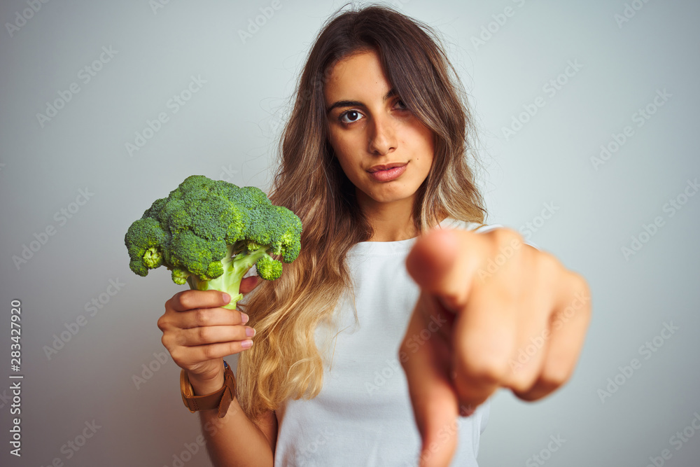 Young beautiful woman eating broccoli over grey isolated background pointing with finger to the camera and to you, hand sign, positive and confident gesture from the front Young beautiful woman eating broccoli over grey isolated background pointing with finger to the camera and to you, hand sign, positive and confident gesture from the front