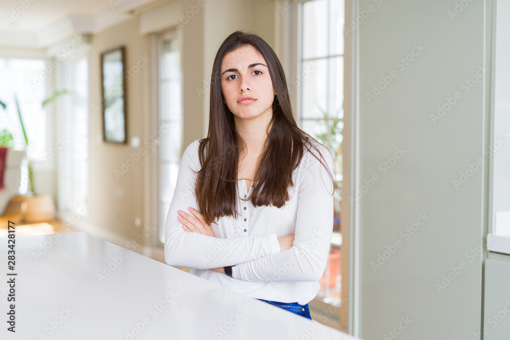 Beautiful young woman sitting on white table at home skeptic and nervous, disapproving expression on face with crossed arms. Negative person. Beautiful young woman sitting on white table at home skeptic and nervous, disapproving expression on face with crossed arms. Negative person.