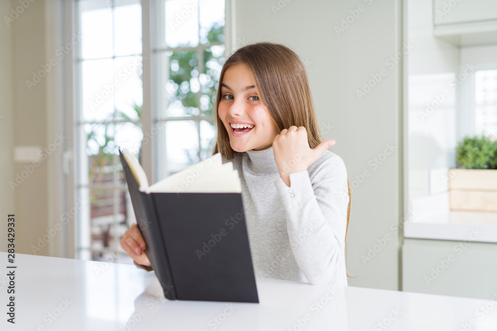 Beautiful young girl reading a book at home pointing and showing with thumb up to the side with happy face smiling Beautiful young girl reading a book at home pointing and showing with thumb up to the side with happy face smiling