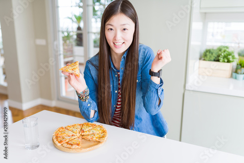 Beautiful Asian woman eating a slice of cheese pizza screaming proud and celebrating victory and success very excited  cheering emotion
