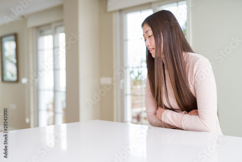 Beautiful Asian woman wearing casual sweater on white table with hand on stomach because nausea, painful disease feeling unwell. Ache concept.