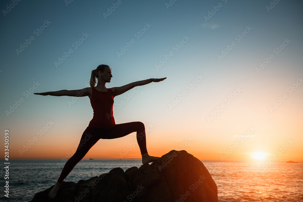 Silhouette of yoga woman doing exercise on the sea beach during amazing surreal sunset.