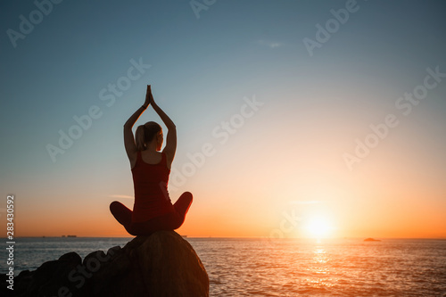 Silhouette of yoga woman doing meditation on the sea beach during amazing surreal sunset.