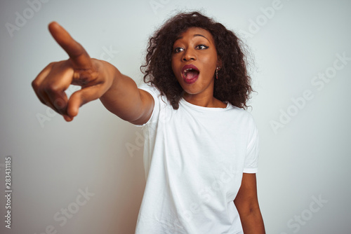 Young african american woman wearing t-shirt standing over isolated white background Pointing with finger surprised ahead, open mouth amazed expression, something on the front