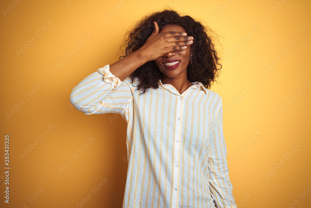 African american woman wearing striped shirt standing over isolated yellow background smiling and laughing with hand on face covering eyes for surprise. Blind concept. African american woman wearing striped shirt standing over isolated yellow background smiling and laughing with hand on face covering eyes for surprise. Blind concept.
