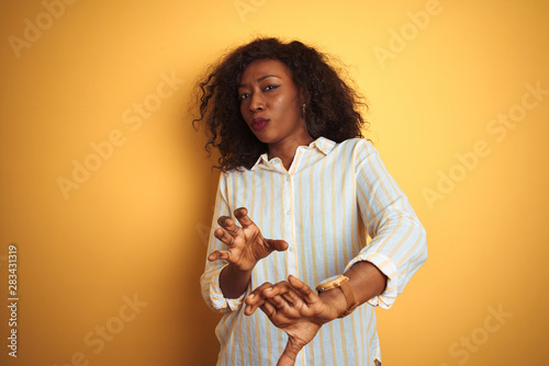 African american woman wearing striped shirt standing over isolated yellow background disgusted expression, displeased and fearful doing disgust face because aversion reaction. With hands raised. 