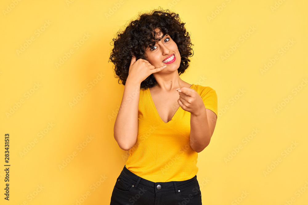 Young arab woman with curly hair wearing t-shirt standing over isolated yellow background smiling doing talking on the telephone gesture and pointing to you. Call me.