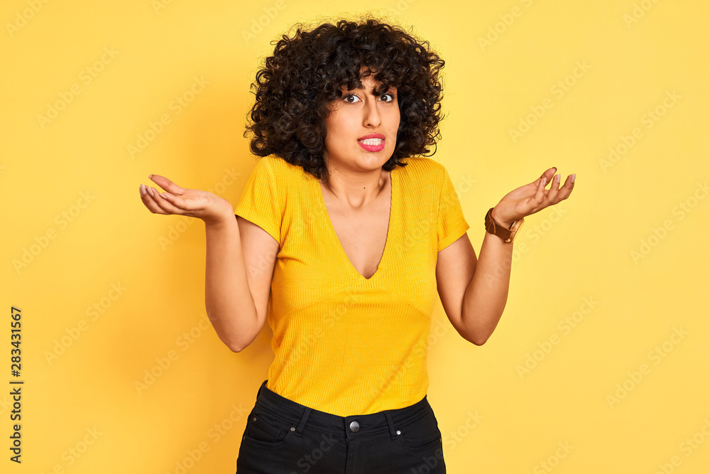Young arab woman with curly hair wearing t-shirt standing over isolated yellow background clueless and confused expression with arms and hands raised. Doubt concept. Young arab woman with curly hair wearing t-shirt standing over isolated yellow background clueless and confused expression with arms and hands raised. Doubt concept.
