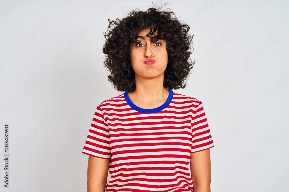 Young arab woman with curly hair wearing striped t-shirt over isolated white background puffing cheeks with funny face. Mouth inflated with air, crazy expression. Young arab woman with curly hair wearing striped t-shirt over isolated white background puffing cheeks with funny face. Mouth inflated with air, crazy expression.