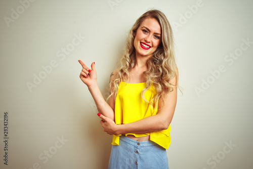 Young beautiful woman wearing yellow t-shirt standing over white isolated background with a big smile on face  pointing with hand and finger to the side looking at the camera.