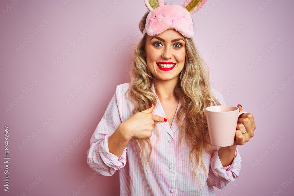 Young woman wearing pajama and mask drinking a cup of coffee over pink isolated background with surprise face pointing finger to himself Young woman wearing pajama and mask drinking a cup of coffee over pink isolated background with surprise face pointing finger to himself
