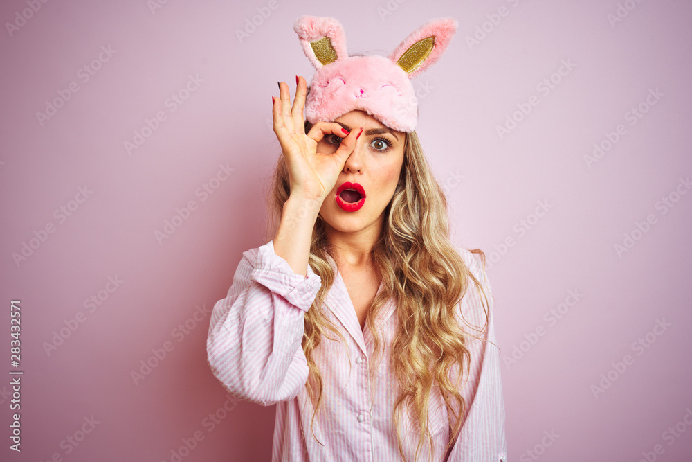 Young beautiful woman wearing pajama and sleep mask over pink isolated background doing ok gesture shocked with surprised face, eye looking through fingers. Unbelieving expression.