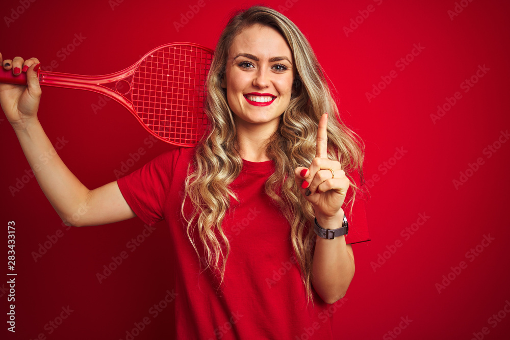 Young sportwoman playing tennis with racket and a ball over red isolated background surprised with an idea or question pointing finger with happy face, number one Young sportwoman playing tennis with racket and a ball over red isolated background surprised with an idea or question pointing finger with happy face, number one
