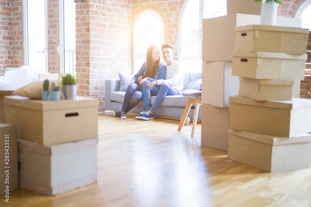 Fototapeta premium Beautiful young asian couple relaxing together sitting on the sofa arround cardboard boxes, happy moving to a new house