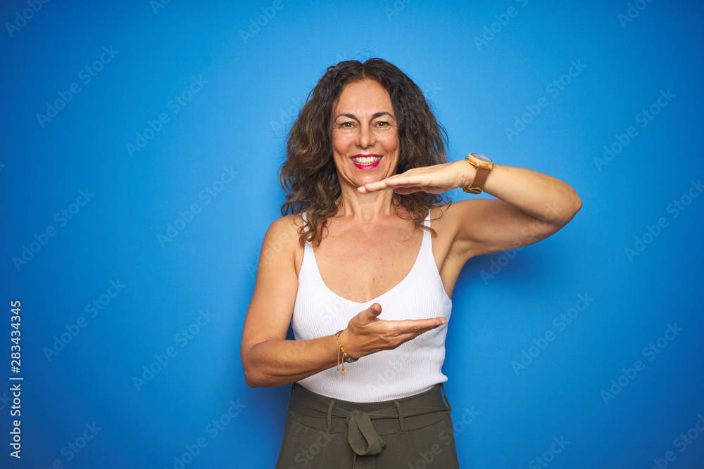 Middle age senior woman with curly hair standing over blue isolated background gesturing with hands showing big and large size sign, measure symbol. Smiling looking at the camera. Measuring concept. Middle age senior woman with curly hair standing over blue isolated background gesturing with hands showing big and large size sign, measure symbol. Smiling looking at the camera. Measuring concept.