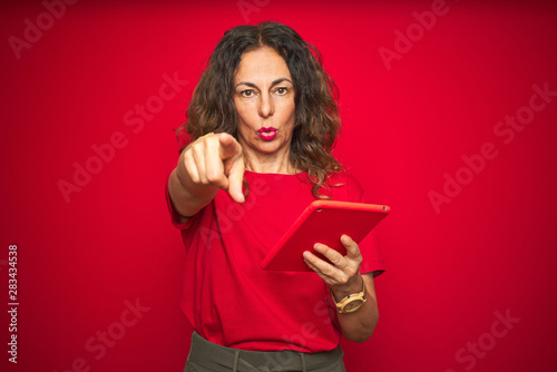 Middle age senior woman using touchpad tablet over red isolated background pointing with finger to the camera and to you, hand sign, positive and confident gesture from the front