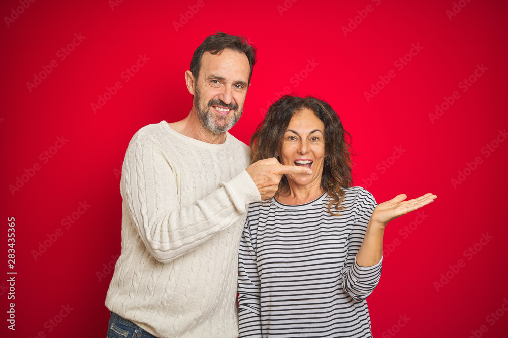 Beautiful middle age couple wearing winter sweater over isolated red background amazed and smiling to the camera while presenting with hand and pointing with finger. Beautiful middle age couple wearing winter sweater over isolated red background amazed and smiling to the camera while presenting with hand and pointing with finger.