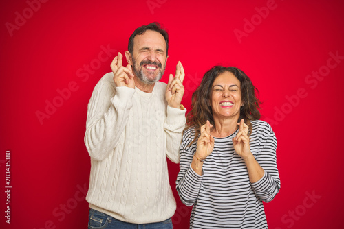 Beautiful middle age couple wearing winter sweater over isolated red background gesturing finger crossed smiling with hope and eyes closed. Luck and superstitious concept.