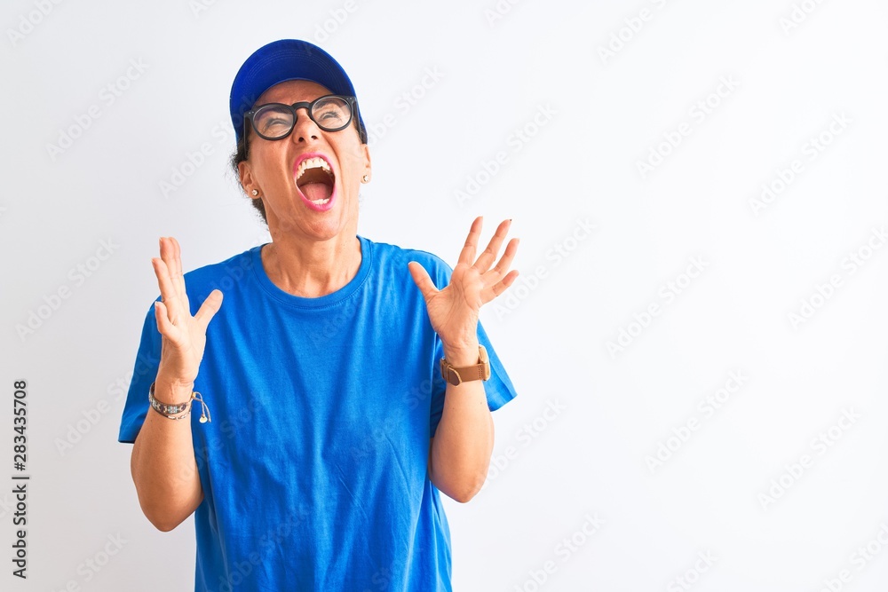 Senior deliverywoman wearing cap and glasses standing over isolated white background crazy and mad shouting and yelling with aggressive expression and arms raised. Frustration concept.