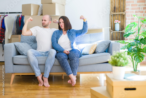 Young couple sitting on the sofa arround cardboard boxes moving to a new house showing arms muscles smiling proud. Fitness concept.